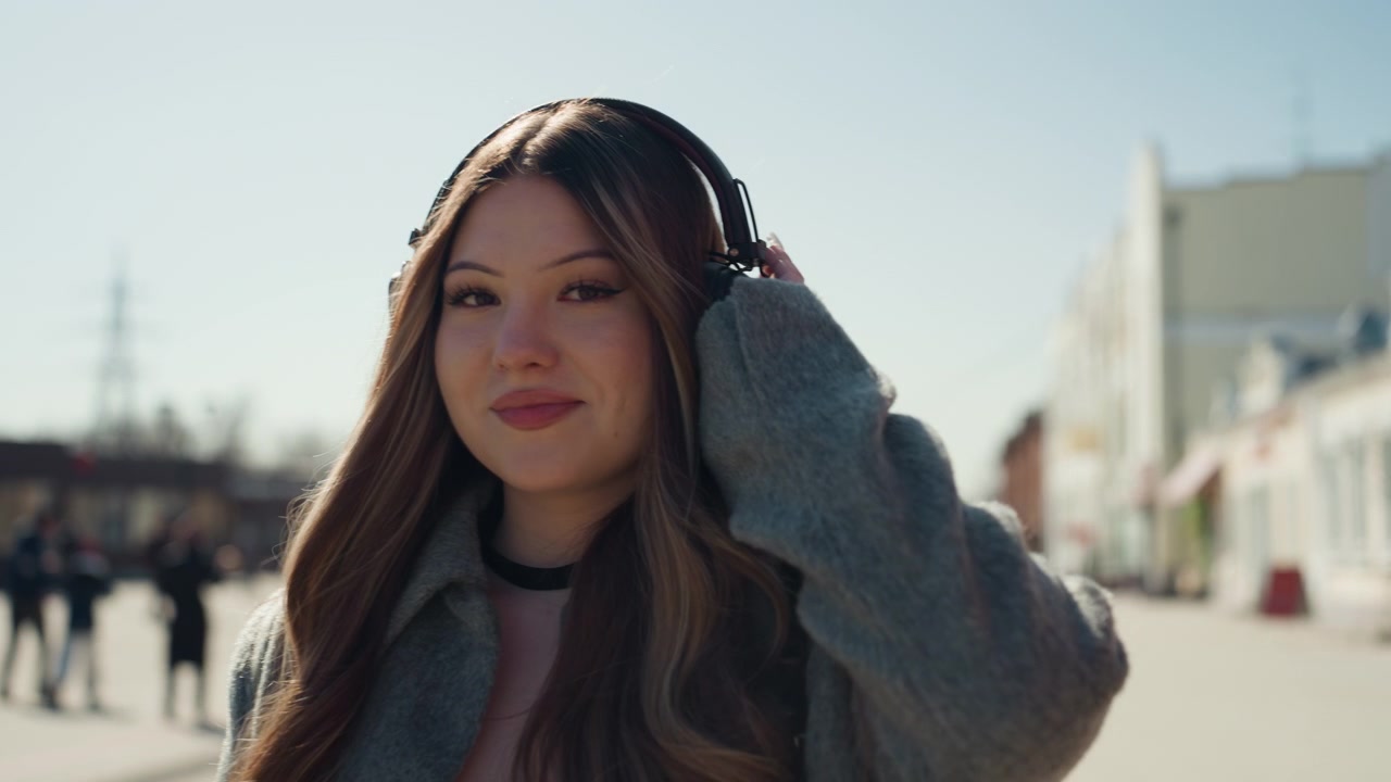 Volto giovane che sogna ad occhi aperti. Ragazza in città che contempla pensieri. Individuo femminile perso in una pacifica rêverie cittadina. Giovane donna rilassata che si gode momenti sereni in mezzo allo scenario cittadino con le cuffie.