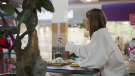 hand inserting straw into coffee cup placed on tray with covered fries in casual dining setting, background includes blurred restaurant elements, chairs, and greenery