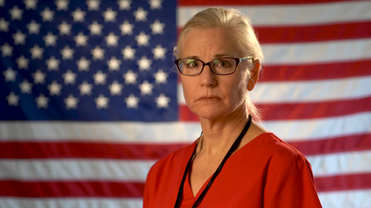 Medium tight portrait of the back of blonde nurses head as she turns and looks at camera looking sad wearing glasses with out of focus US flag
