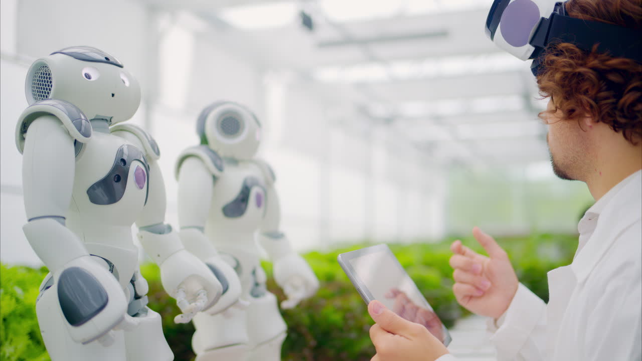 Laboratory technician in a white coat wearing virtual reality headset analysing graphs on a tablet while interacting with two humanoid robots in a greenhouse farm