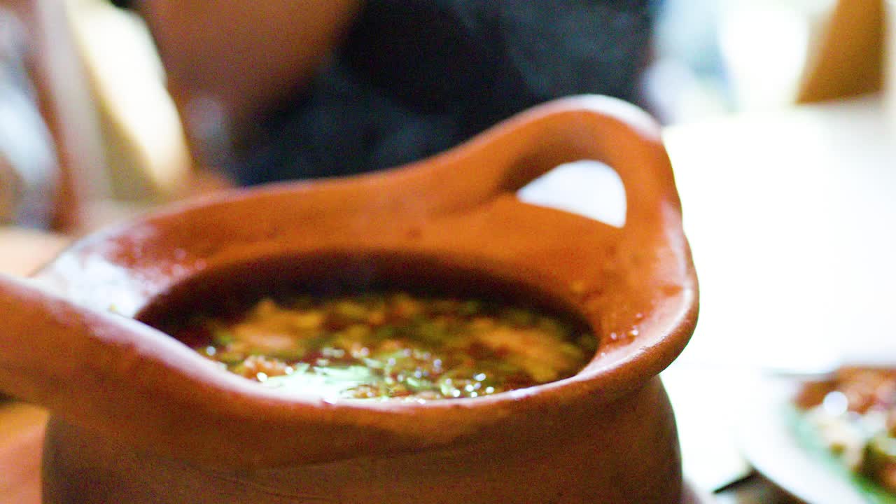 Hand lifts clay pot lid, revealing steaming herbal Thai soup in warm, natural restaurant lighting