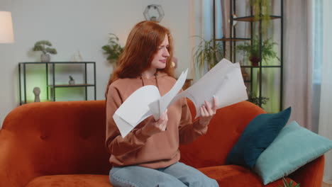 stressed woman freelancer sitting on sofa reviewing charts and graphs and throwing papers in air