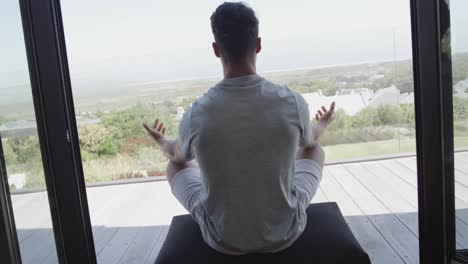 rear view of biracial man sitting on sunny balcony practicing yoga meditation, slow motion