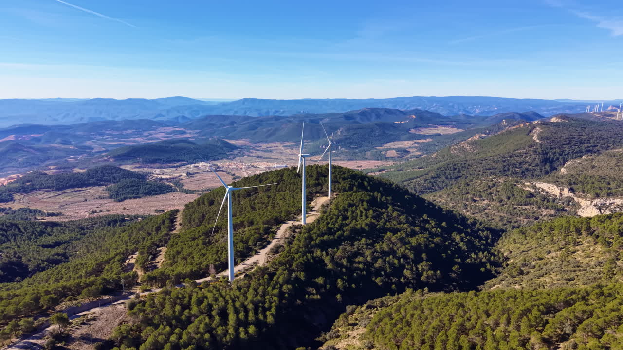 Aerogeneradores inmóviles en una cresta montañosa boscosa con vistas panorámicas del valle y la cordillera bajo un cielo azul claro