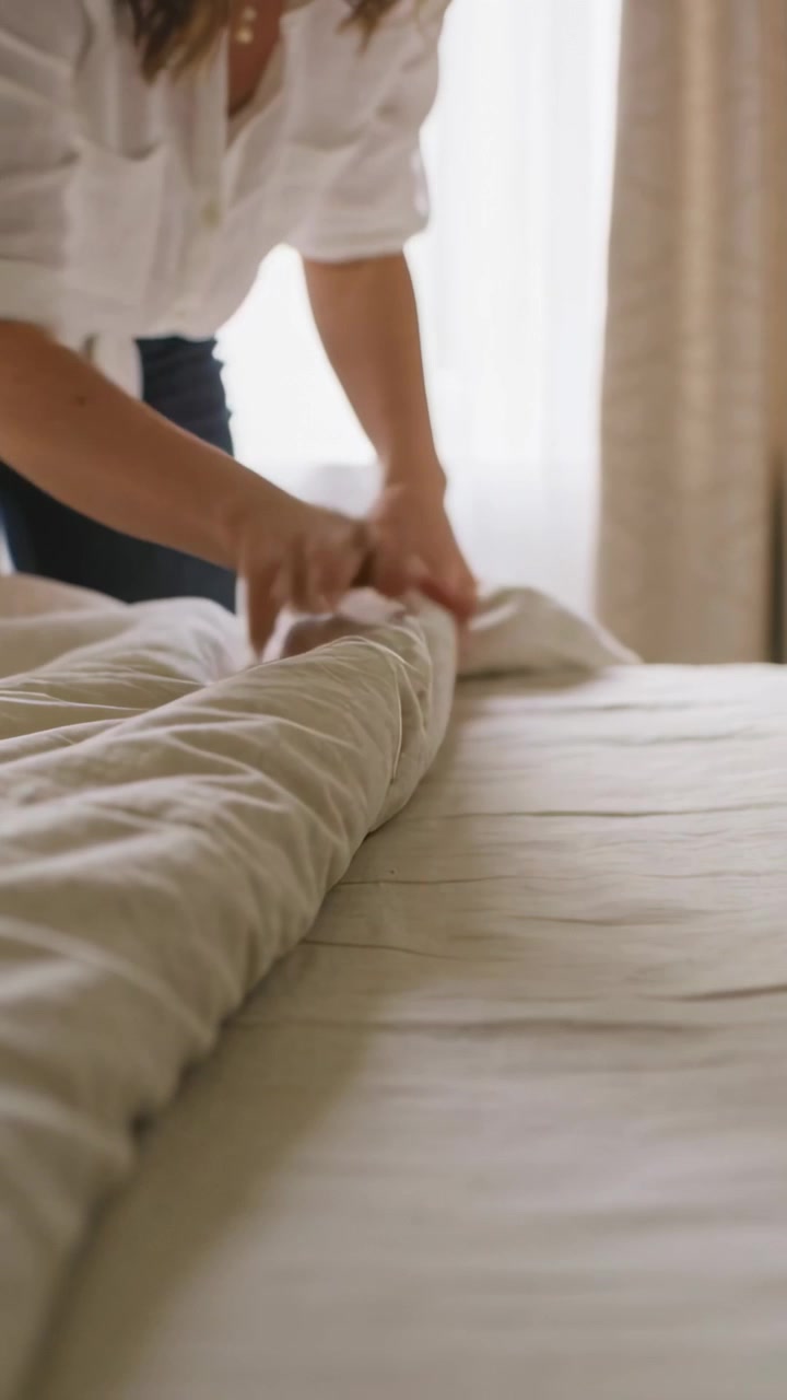 Video vertical: Mujer con camisa blanca sujetando el edredón, arreglando y alisando la cama en casa para descansar.