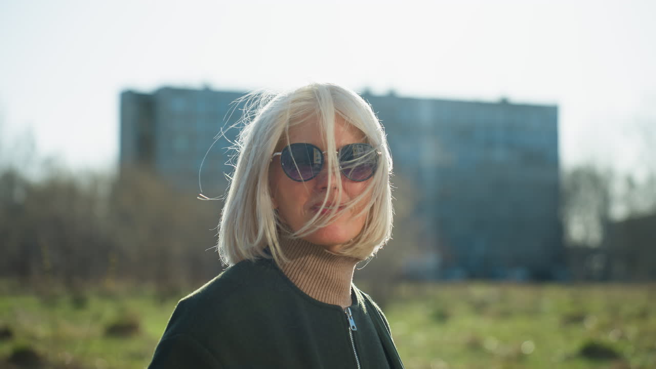 Elegante donna bionda con taglio bob che indossa occhiali da sole e un cappotto scuro, in piedi all'aperto in un contesto urbano, con il vento che le soffia i capelli sul viso e la luce del sole che crea un'atmosfera suggestiva con edifici cittadini sfocati