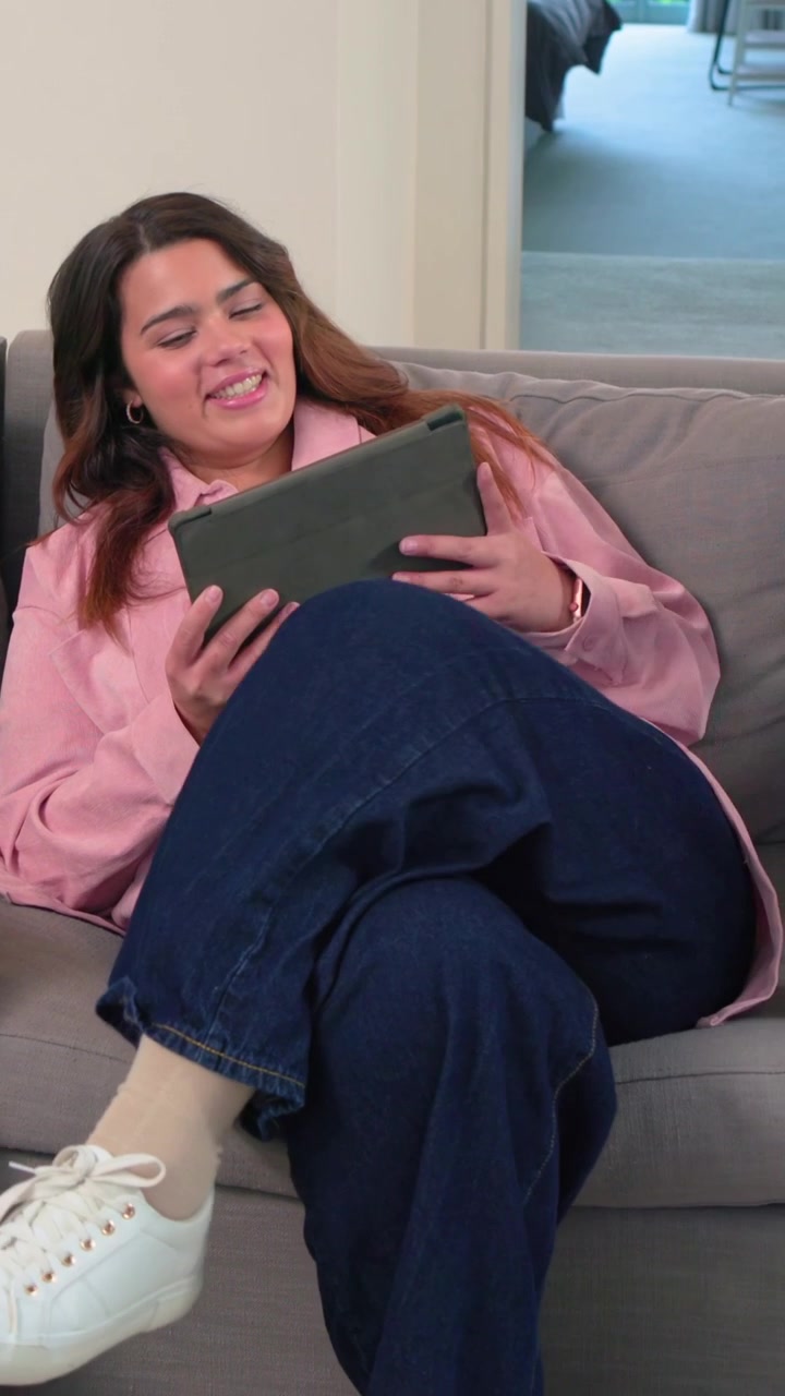 Video verticale: Donna adulta che nota un tablet, regola e tocca lo schermo su un divano grigio, leggendo.