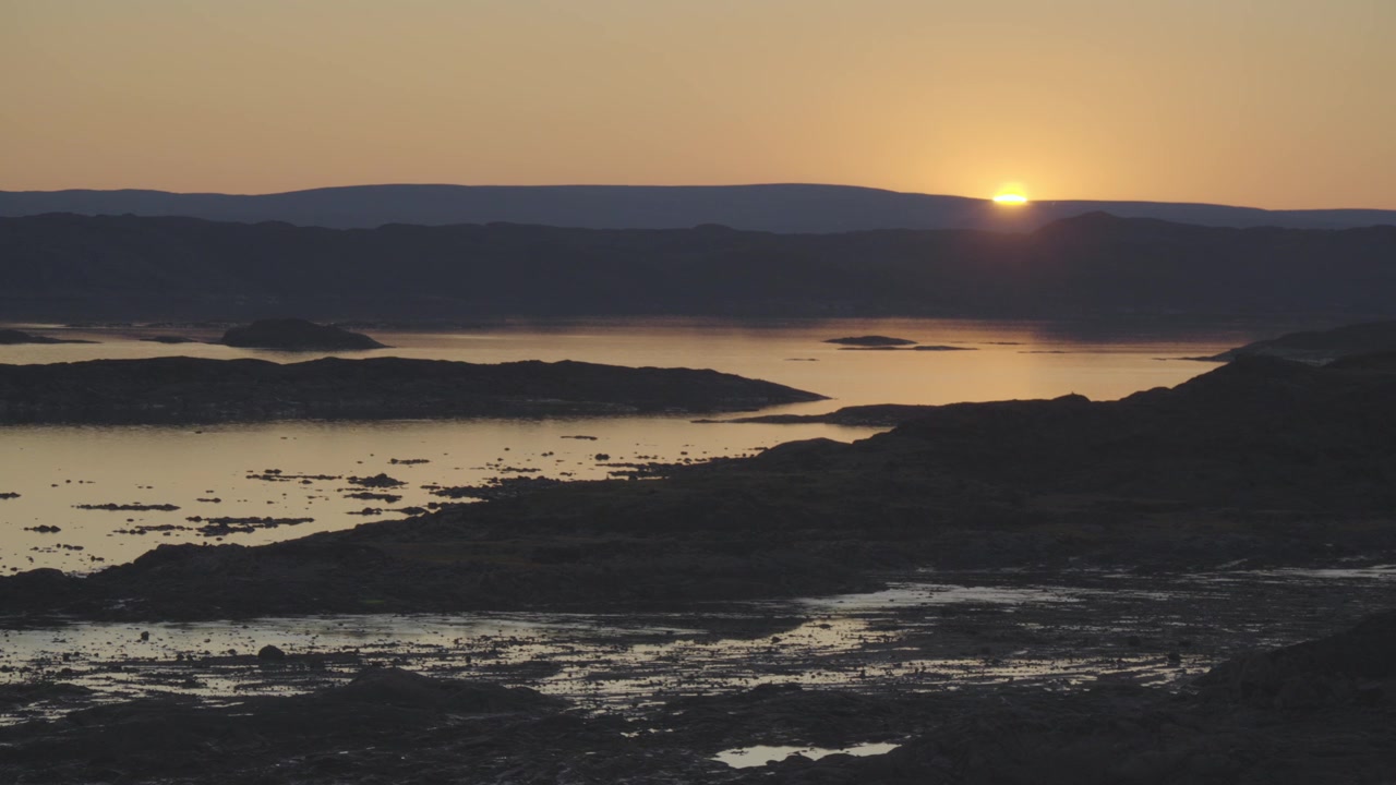 Clipe contínuo de paisagem em tempo real de 30 segundos capturando o sol ártico mergulhando lentamente abaixo de colinas distantes da tundra, transformando o céu e a água em dourado sobre a Baía de Frobisher.