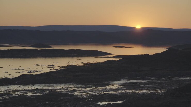 Clipe contínuo de paisagem em tempo real de 30 segundos capturando o sol ártico mergulhando lentamente abaixo de colinas distantes da tundra, transformando o céu e a água em dourado sobre a Baía de Frobisher.