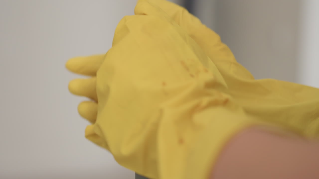 Close up detailed view of cleaner hands in yellow gloves gripping mop handle, one hand placed over the other, emphasizing protective gloves and household cleaning task with blurred background