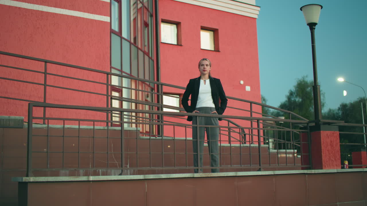 Un lavoratore da remoto posa con sicurezza, mani in tasca, fuori dalla sua residenza con muro rosso, alberi sullo sfondo, lampione acceso al tramonto, ringhiera e muro piastrellato che riflettono la luce ambientale nelle vicinanze