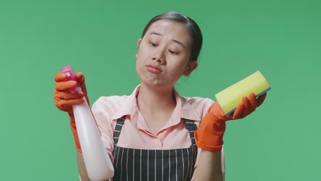 un primer plano de una ama de casa asiática desconcertada con un delantal que muestra un spray y una esponja de cocina mientras está de pie en el estudio de pantalla verde