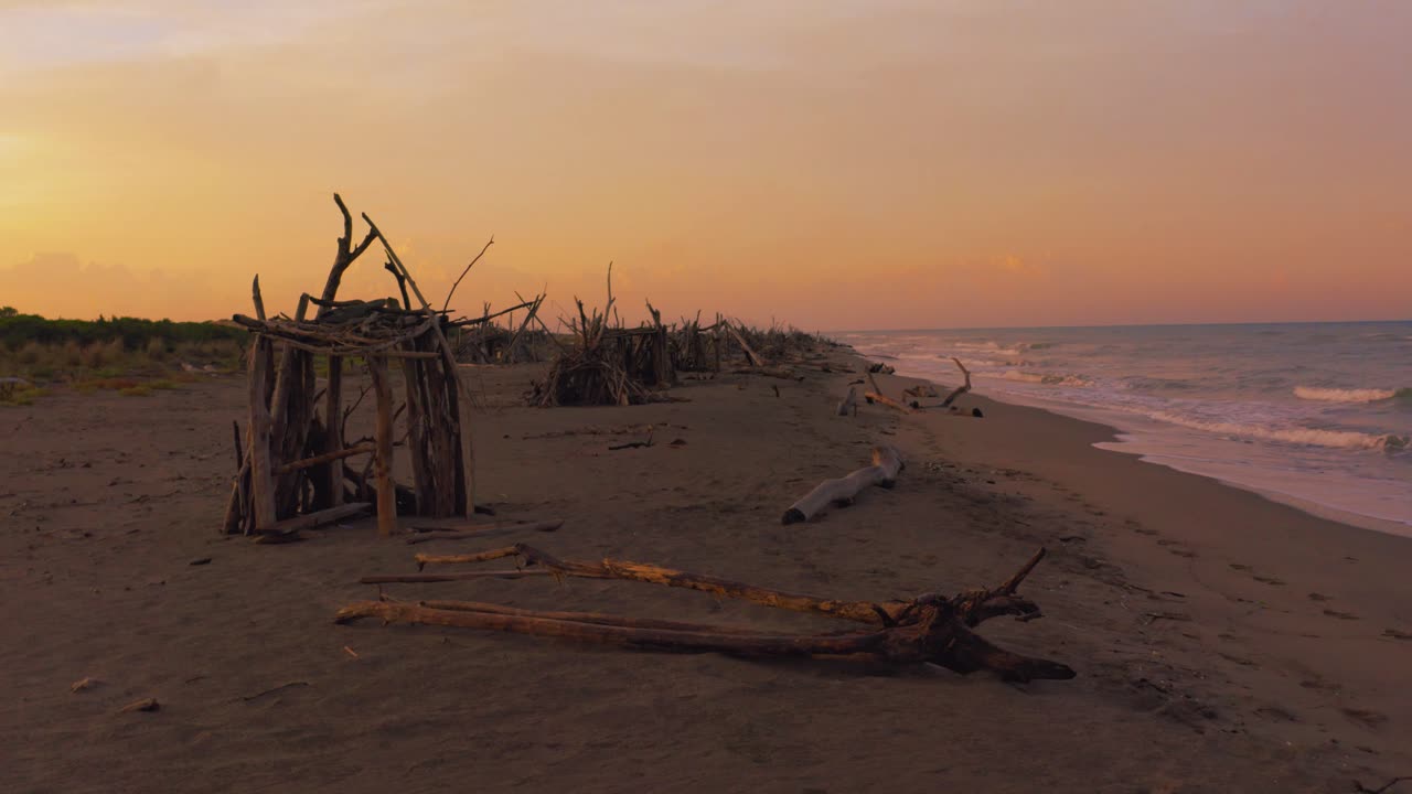 imágenes aéreas de drones de hermosos tipis de madera a la deriva en una playa de arena junto al mar en el icónico parque natural maremma en toscana, italia, con un espectacular cielo de nubes rojas al atardecer con pequeñas olas azules