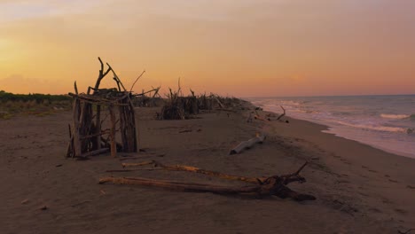 aerial drone footage of beautiful driftwood tipis at a sandy beach seaside in the iconic maremma nature park in tuscany, italy, with a dramatic red cloud sky at sunset with small blue waves