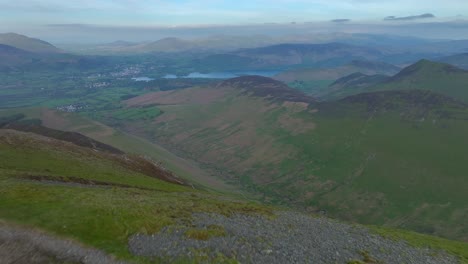 Mountain-walker-on-summit-with-flyover-revealing-deep-valley,-lowlands-and-lake-with-distant-mountains-behind,-prior-to-sunset