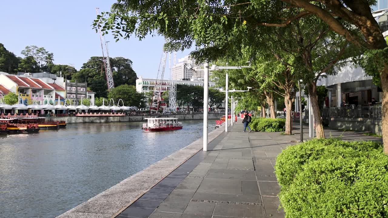 A tourist boat glides along a riverside promenade lined with trees and colorful buildings in bright daylight, captured in a steady wide shot