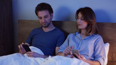 caucasian couple sitting on the bed and using smartphones at night