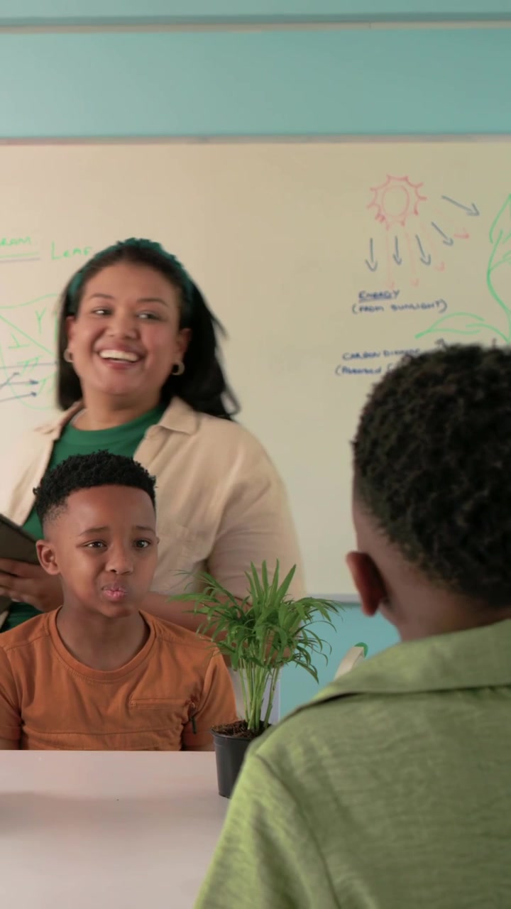 Vertical video: Tapping tablet, teacher in green engaging kids at counter with whiteboard and plant
