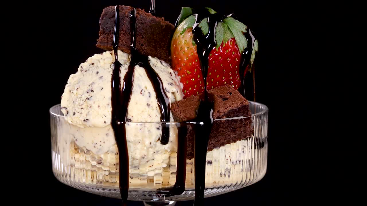 A close-up of vanilla ice cream with chocolate chips, brownies, and strawberries as chocolate sauce is poured over, set against a black background with dramatic lighting