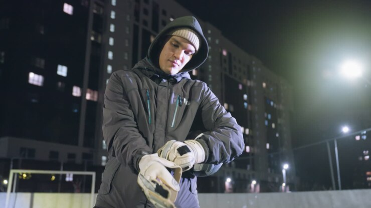 Goalkeeper adjusts gloves with ball securely tucked under arm during evening practice session on urban field, focused expression and illuminated background
