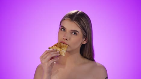 static shot of a beautiful young woman eating a slice of delicious margherita pizza while taking the first bite and looking contentedly at the camera in front of purple background in slow motion