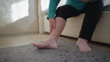 old woman in green applying ointment to her leg while sitting on bed. one sock on, the other off, in a peaceful bedroom setting. soft light fills the room, creating a calm and reflective atmosphere