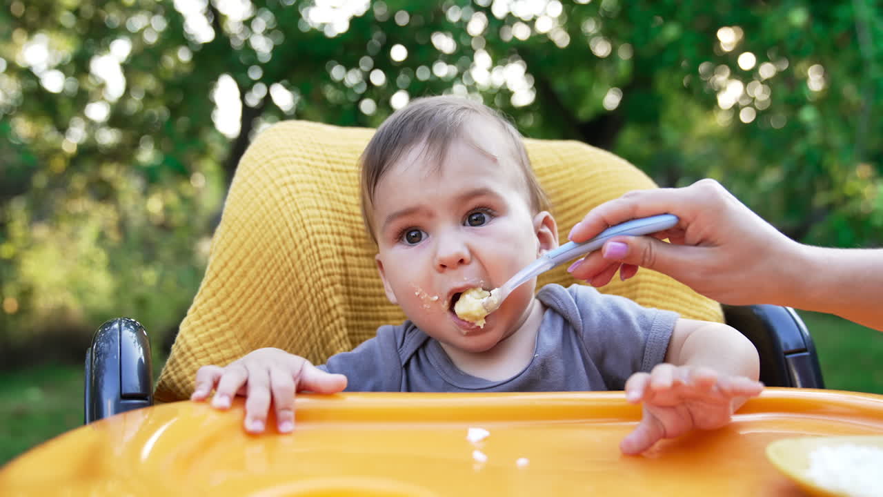 Lovely sweet boy nutrition outdoors. Mother gives a spoon with food to her little son. Blurred nature background.