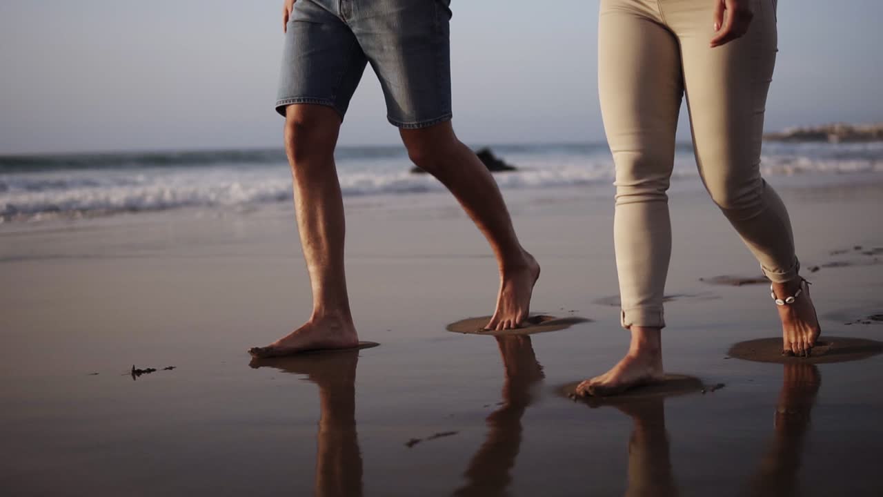 pareja de jóvenes irreconocibles caminando juntos en la arena dorada de la playa. piernas masculinas y femeninas caminando cerca del océano. pies descalzos de pareja yendo en la playa de arena con olas. vacaciones de verano