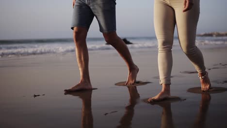 Unrecognizable-young-couple-stepping-together-at-the-golden-sand-at-sea-beach.-Male-and-female-legs-walking-near-ocean.-Bare-feet-of-pair-going-on-sandy-shore-with-waves.-Summer-vacation