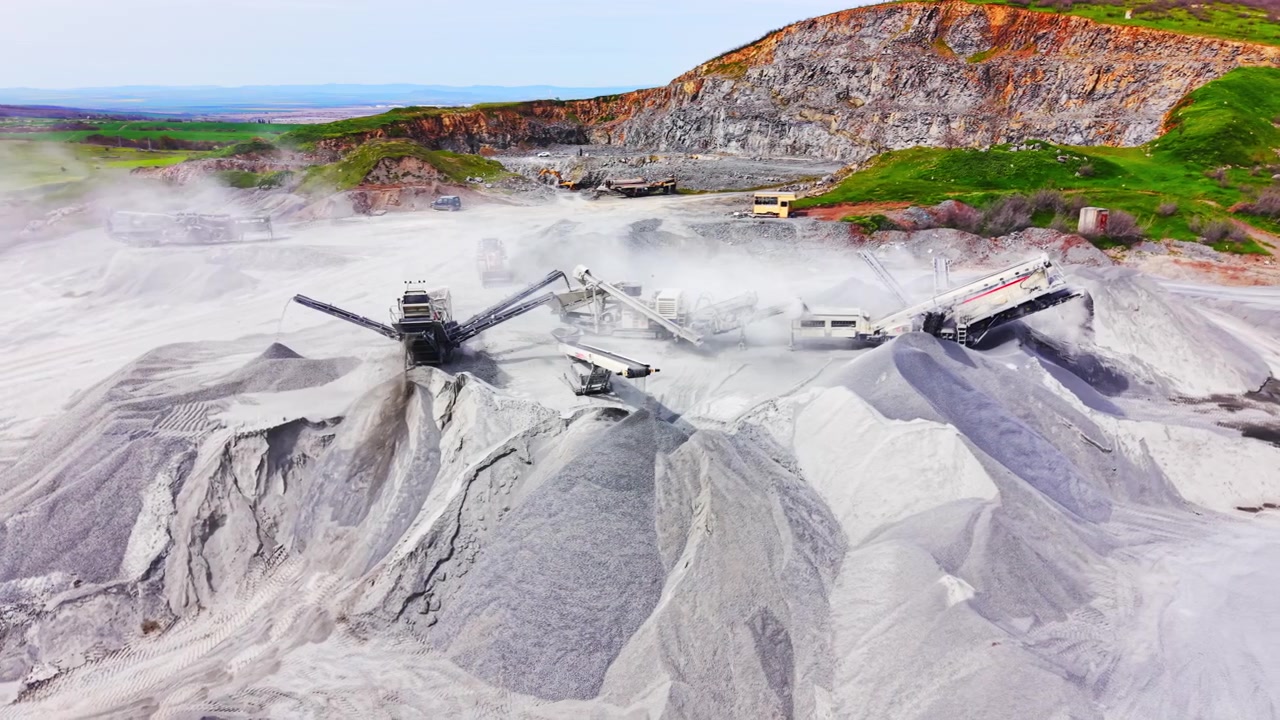 Mobile crushers operating below rocky quarry wall. Dust clouds, gravel mounds and colorful hillside vegetation.