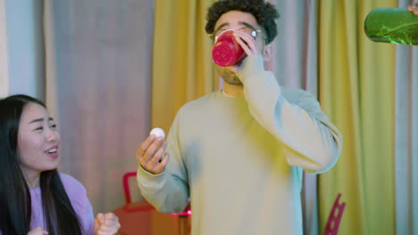 happy multiethnic young friends playing beer pong at home. caucasian boy drinking beer from a plastic cup.