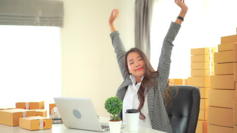 A-young-working-woman-sitting-at-a-desk-surrounded-by-mailing-boxes-reaches-up-to-stretch