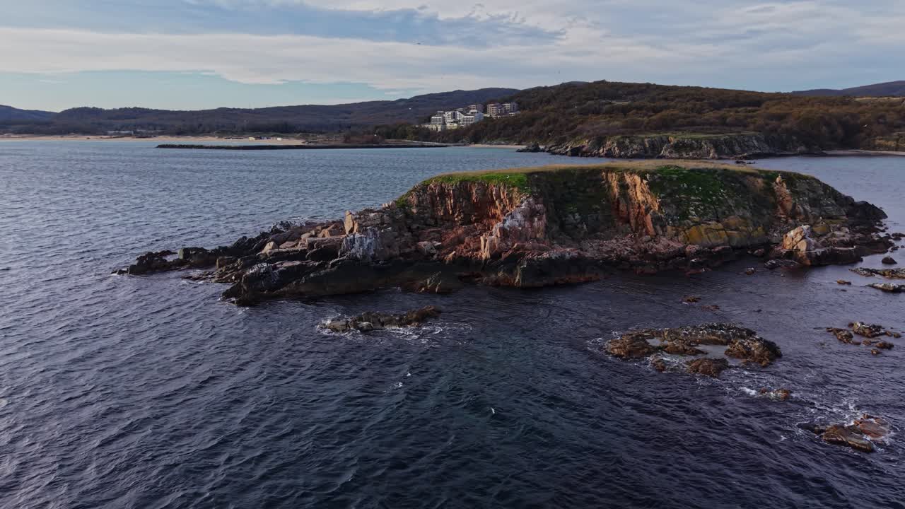 Coastal aerial view of rocky island near serene waters and distant homes