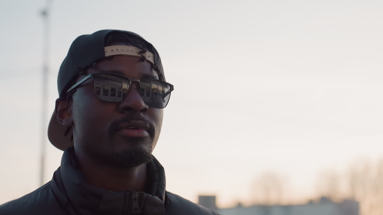 Close up of stylish man wearing sunglasses and backward cap walking outdoors during sunset, with reflection of modern building visible in lenses, exuding confident and relaxed urban vibe