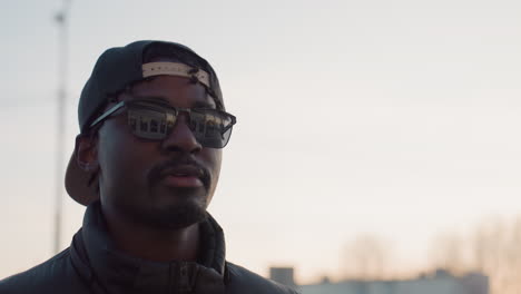 close up of stylish man wearing sunglasses and backward cap walking outdoors during sunset, with reflection of modern building visible in lenses, exuding confident and relaxed urban vibe