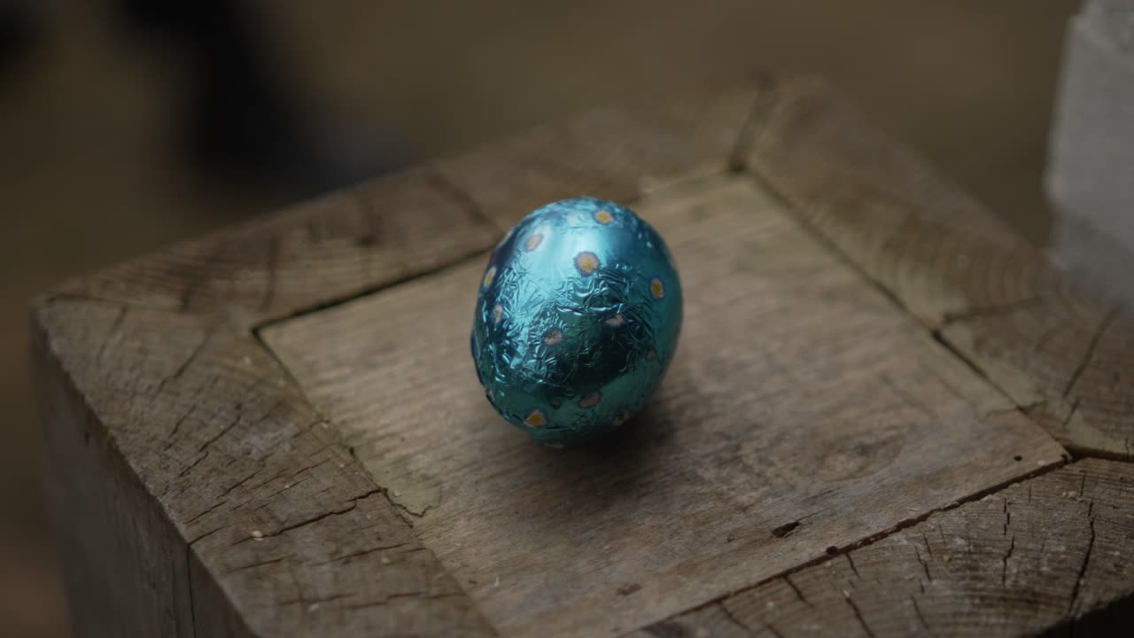 Blue foil-wrapped chocolate Easter egg with yellow polka dots sits on rustic, weathered wooden surface, emphasizing cozy, natural atmosphere. Simple setup highlights contrast of colors and textures.