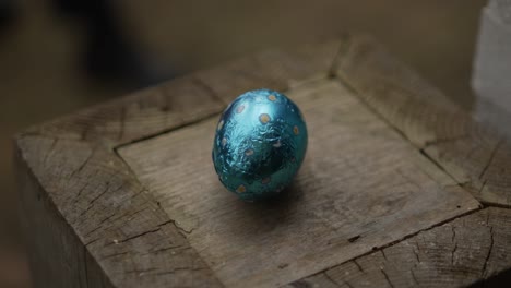 blue foil-wrapped chocolate easter egg with yellow polka dots sits on rustic, weathered wooden surface, emphasizing cozy, natural atmosphere. simple setup highlights contrast of colors and textures.
