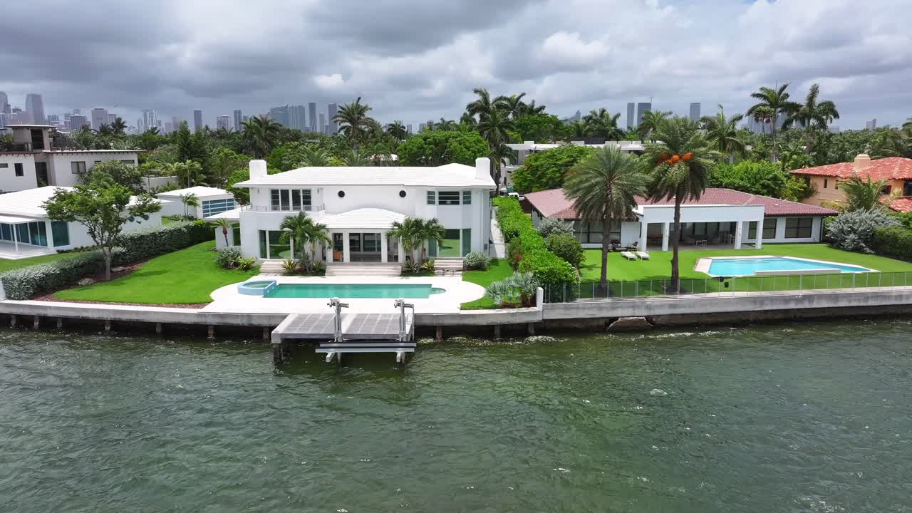 Waterfront mansions Miami Beach, featuring modern architecture, pools, palm trees, and the Miami skyline in the background. Low aerial truck shot over Biscayne Bay. Venetian Islands.