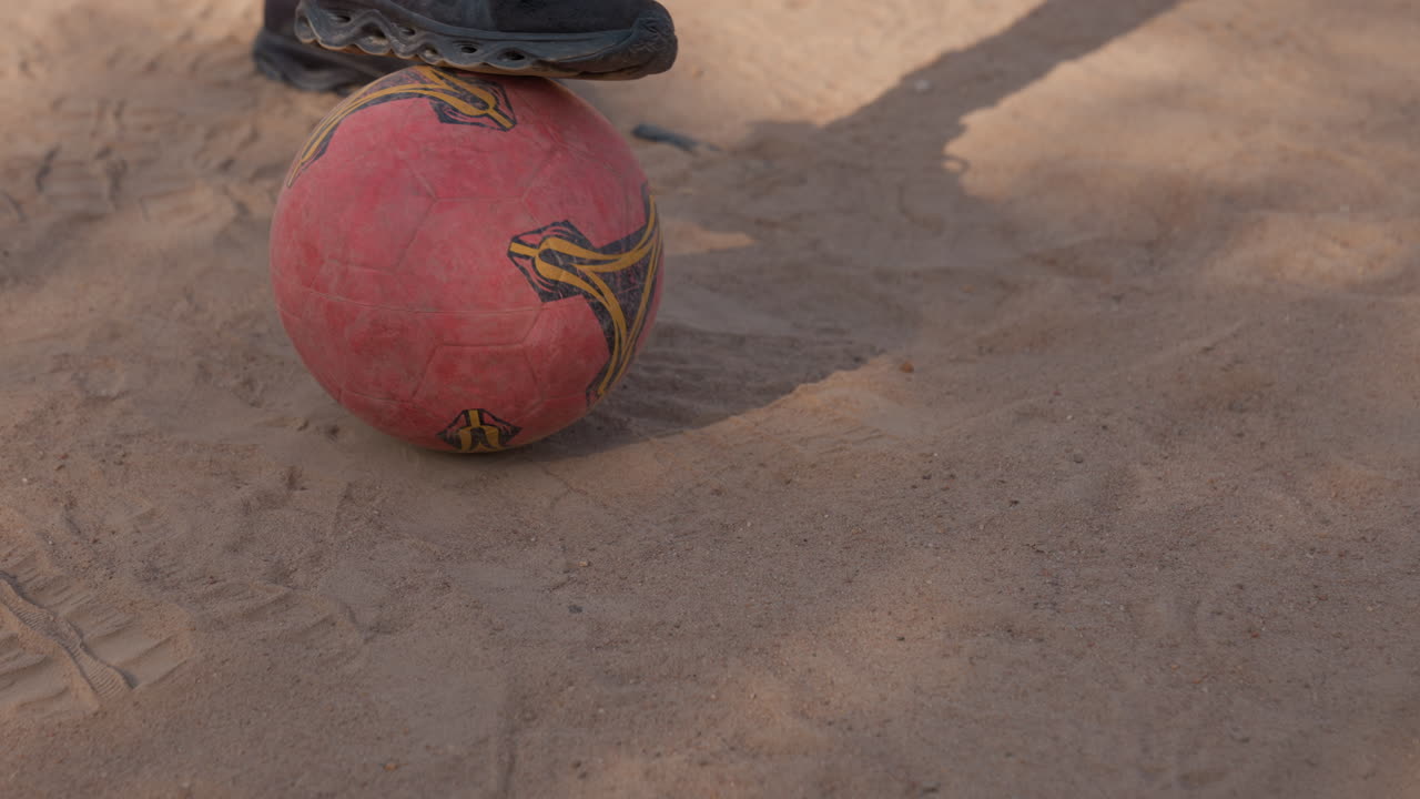 Pie de atleta negro presionando un balón rojo contra el suelo, primer plano detallado de la huella del zapato en un balón de goma desgastado, superficie polvorienta, sombra sutil y contraste, presión cuidadosa y equilibrio, barrio a mediodía.