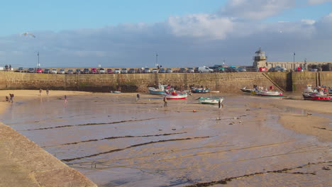 People-Walking-On-Low-Tide-Shore-During-Summertime-Background-With-Moored-Fishing-Boats,-Lighthouse,-And-Seawall-In-St