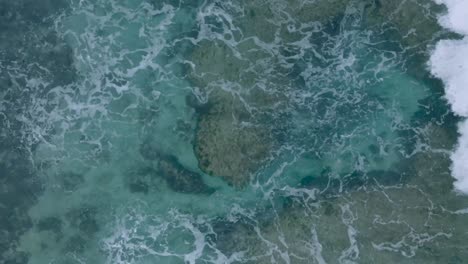 Top-Down-Slow-Motion-Drone-View-of-turquoise-clear-water-and-crashing-waves-over-shallow-coral-reef-in-Uluwatu-Bali-Indonesia