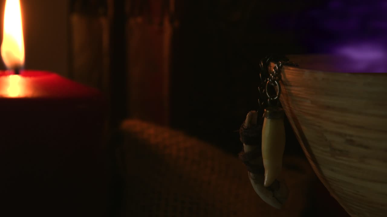 Dettaglio di una stanza di strega dove si preparano pozioni, con vapore viola che esce da una ciotola di legno da cui pendono denti di animale, accanto a una candela con una fiamma tremolante e vecchi libri sullo sfondo.