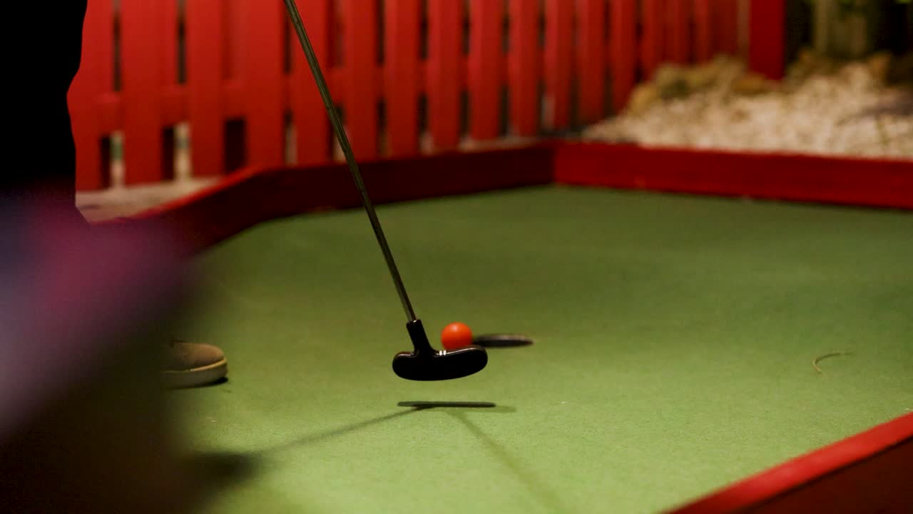 A putter hits a golf ball on a mini-golf course with colorful barriers under warm lighting