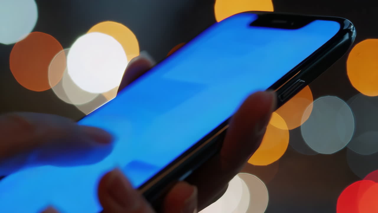 Close-up of hands using a smartphone with a bright blue screen against a background of blurred city lights at night