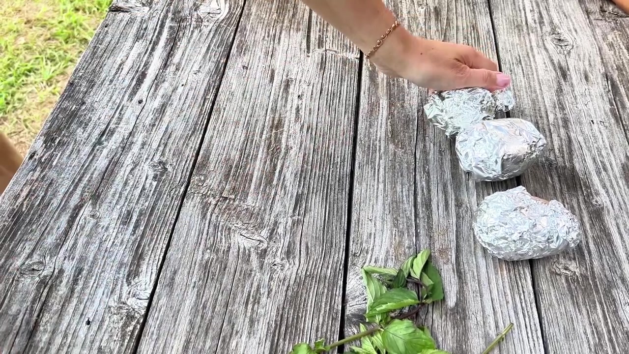 A person wraps whole raw potatoes in aluminum foil on a wooden table. Suitable for food preparation tutorials, outdoor cooking guides, and summer barbecue content.