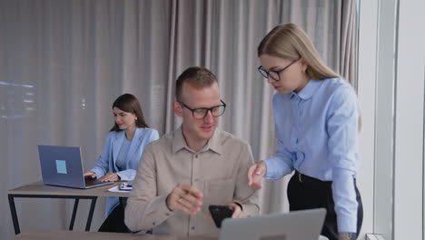 people are busy with their work in the office. man and woman discussing some job issues looking at phone. brunette colleague works on laptop at desk behind.