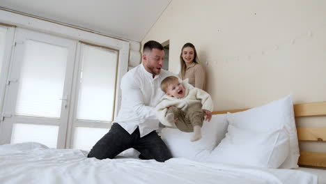 loving caucasian father waves his laughing baby boy on the bed. dad throws his kid into the pillows. mother at backdrop smiles.