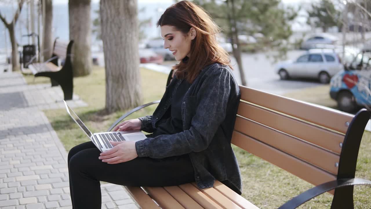 Attractive female in her 20's wearing hipster urban outfit sitting on the bench and typing something on her laptop keyboard