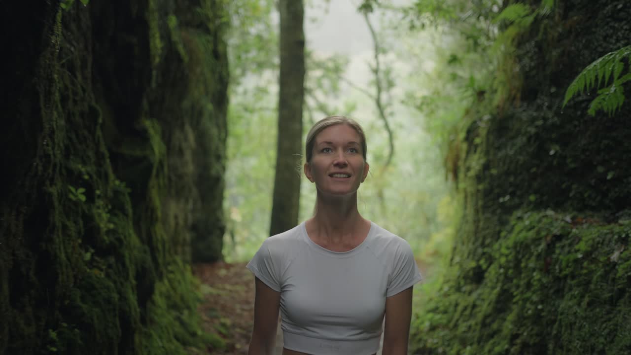 mujer joven con camisa blanca camina a través del paso de montaña del cañón cubierto de musgo, sonriendo y mirando