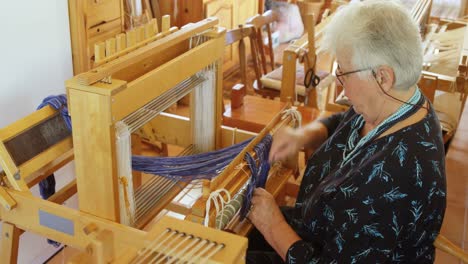 Side-view-of-old-caucasian-senior-woman-preparing-and-sitting-at-hand-loom-machine-in-a-workshop-4k